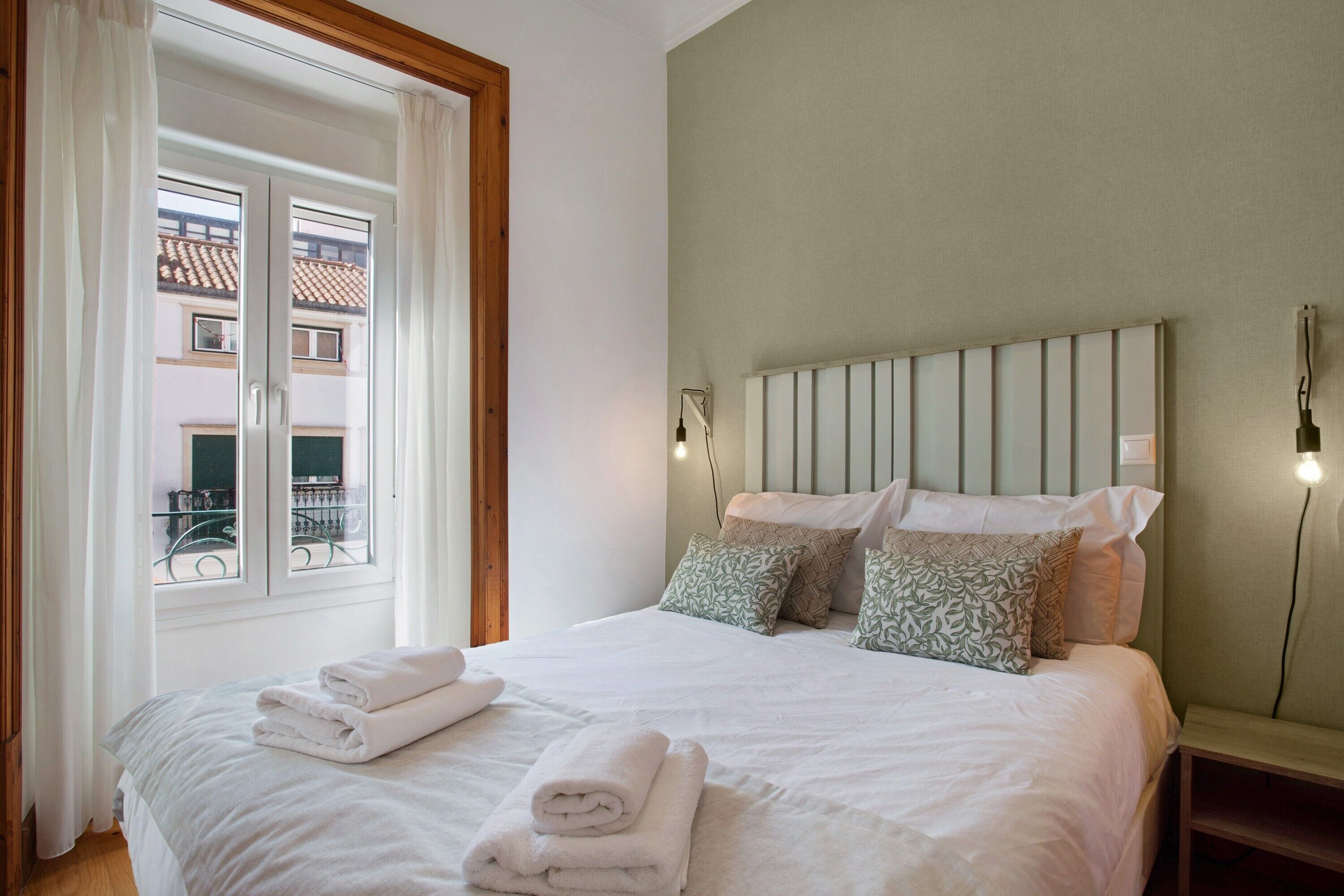 A bedroom with a bed, pillows, a nightstand, a window with curtains, and a view of buildings outside.