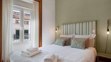 Een slaapkamer met een bed, kussens, een nachtkastje, een raam met gordijnen en uitzicht op gebouwen.