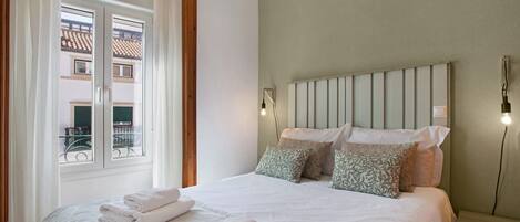A bedroom with a bed, pillows, a nightstand, a window with curtains, and a view of buildings outside.