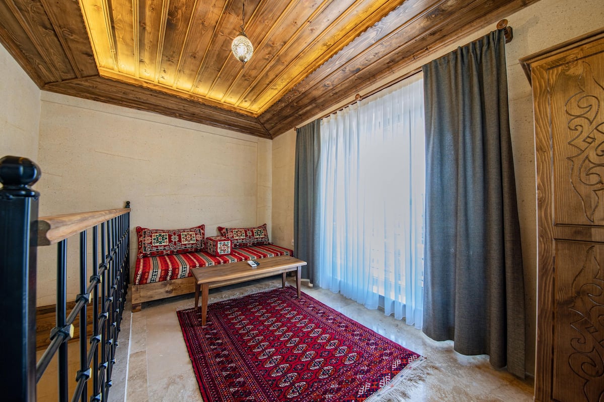 Una habitación con techo de madera, un banco con cojines estampados, una alfombra roja y un ventanal con cortinas.