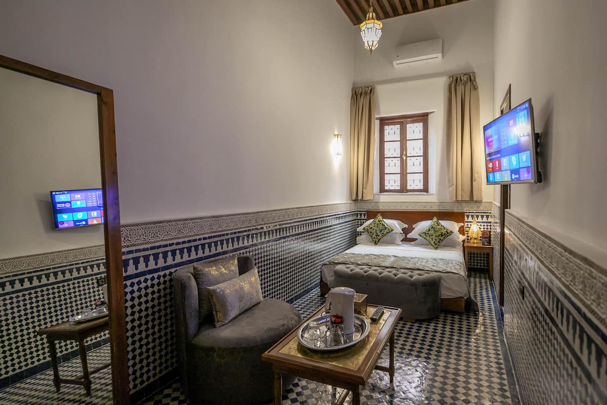 A room with a checkered floor, a bed with decorative pillows, a small table with a tray, a television on the wall, and a window with curtains.
