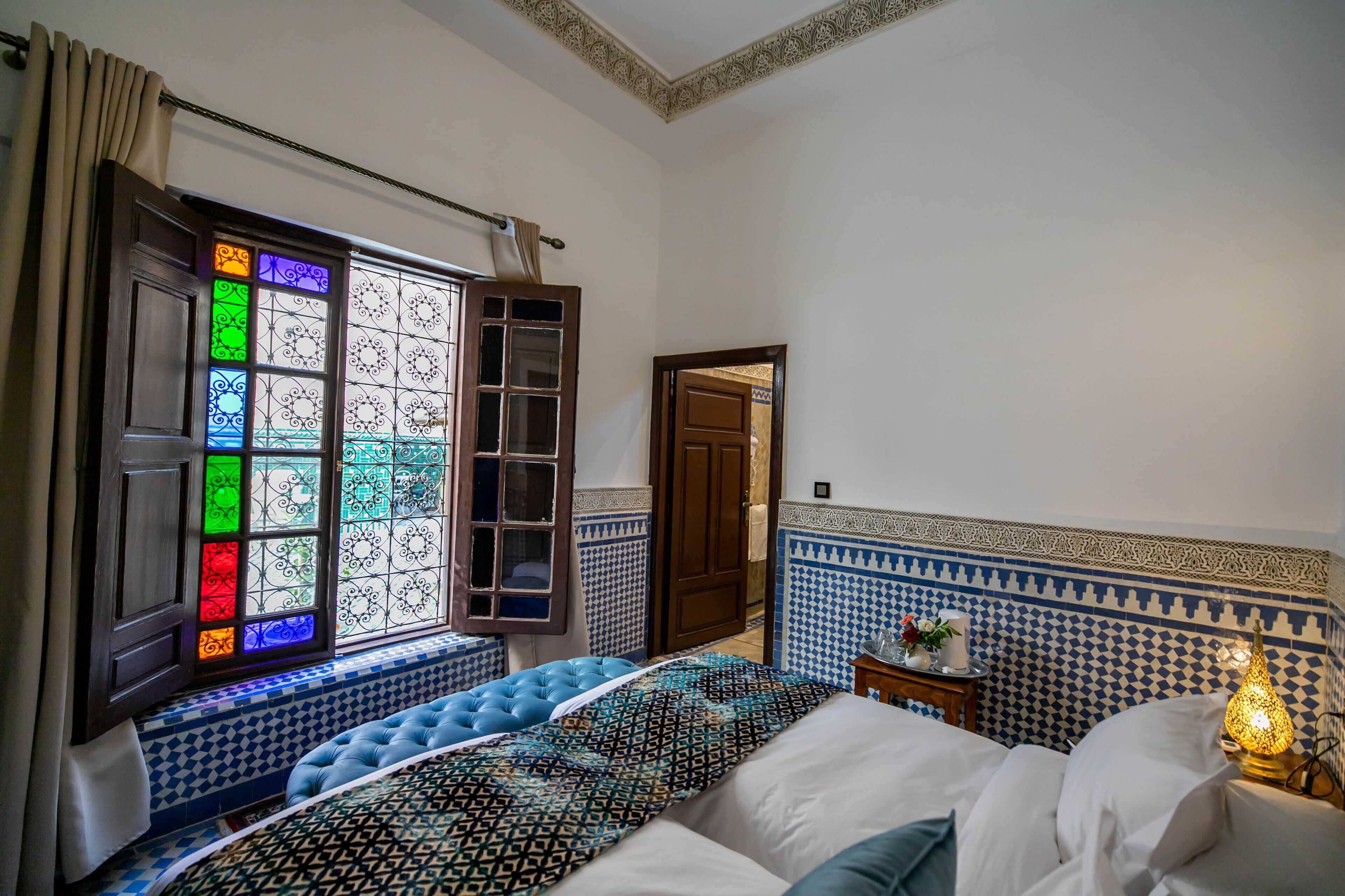 Een slaapkamer met een betegelde muur met patroon, een bed met een beddegoed met patroon, een raam met glas-in-lood en een nachtkastje met een lamp.
