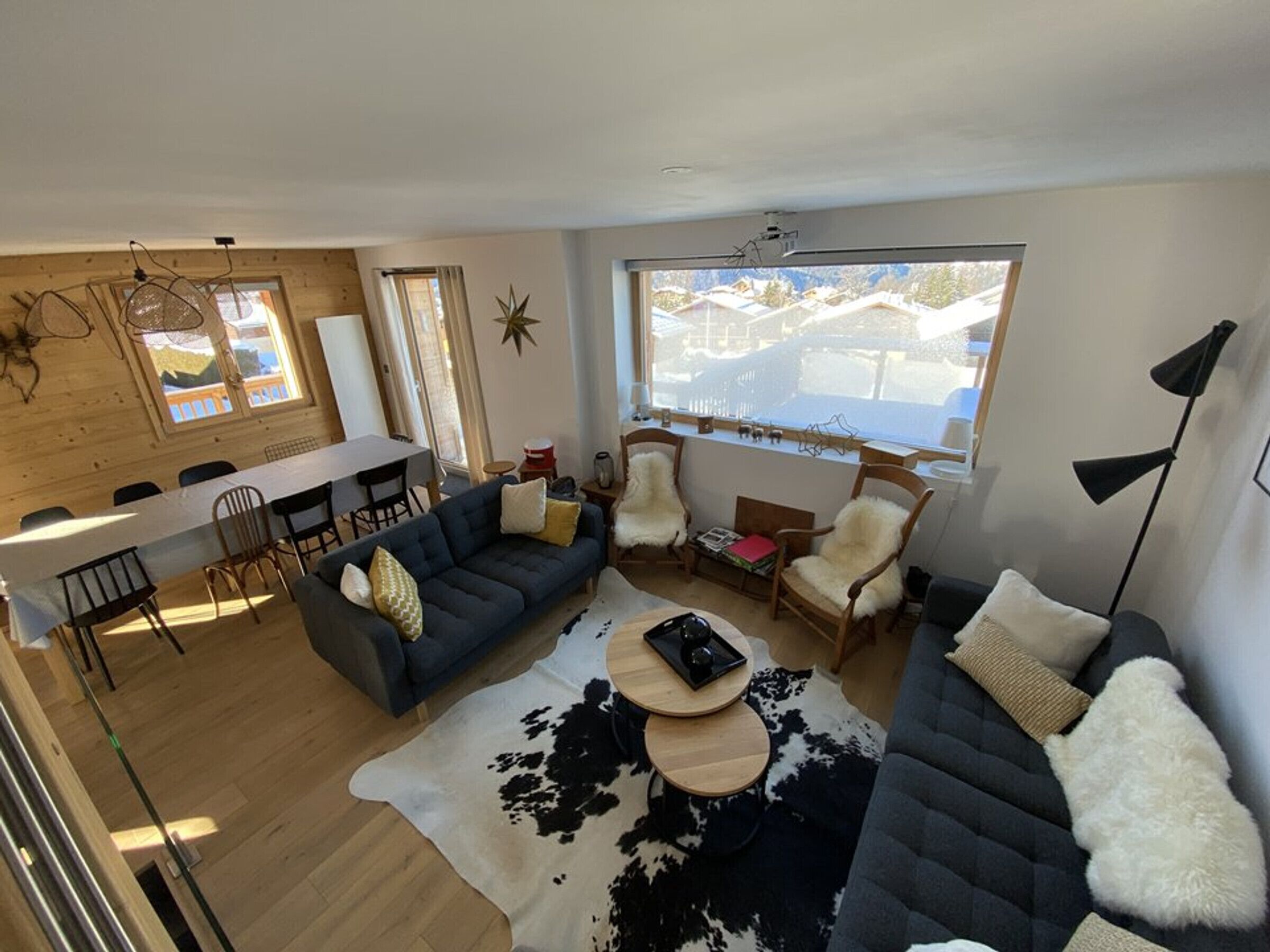 A modern living room with a sofa, chairs, a dining table, and a large window with a view of houses.