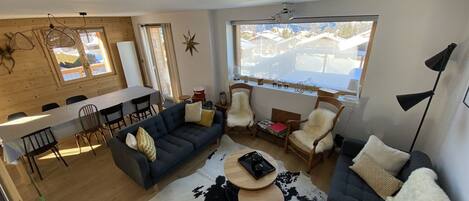 A modern living room with a sofa, chairs, a dining table, and a large window with a view of houses.