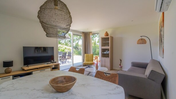 A modern living room with a marble dining table, a sofa, a television, and a large window. - What immediately catches your eye upon entering is the garden-oriented living room with open kitchen (Burgh-Haamstede)