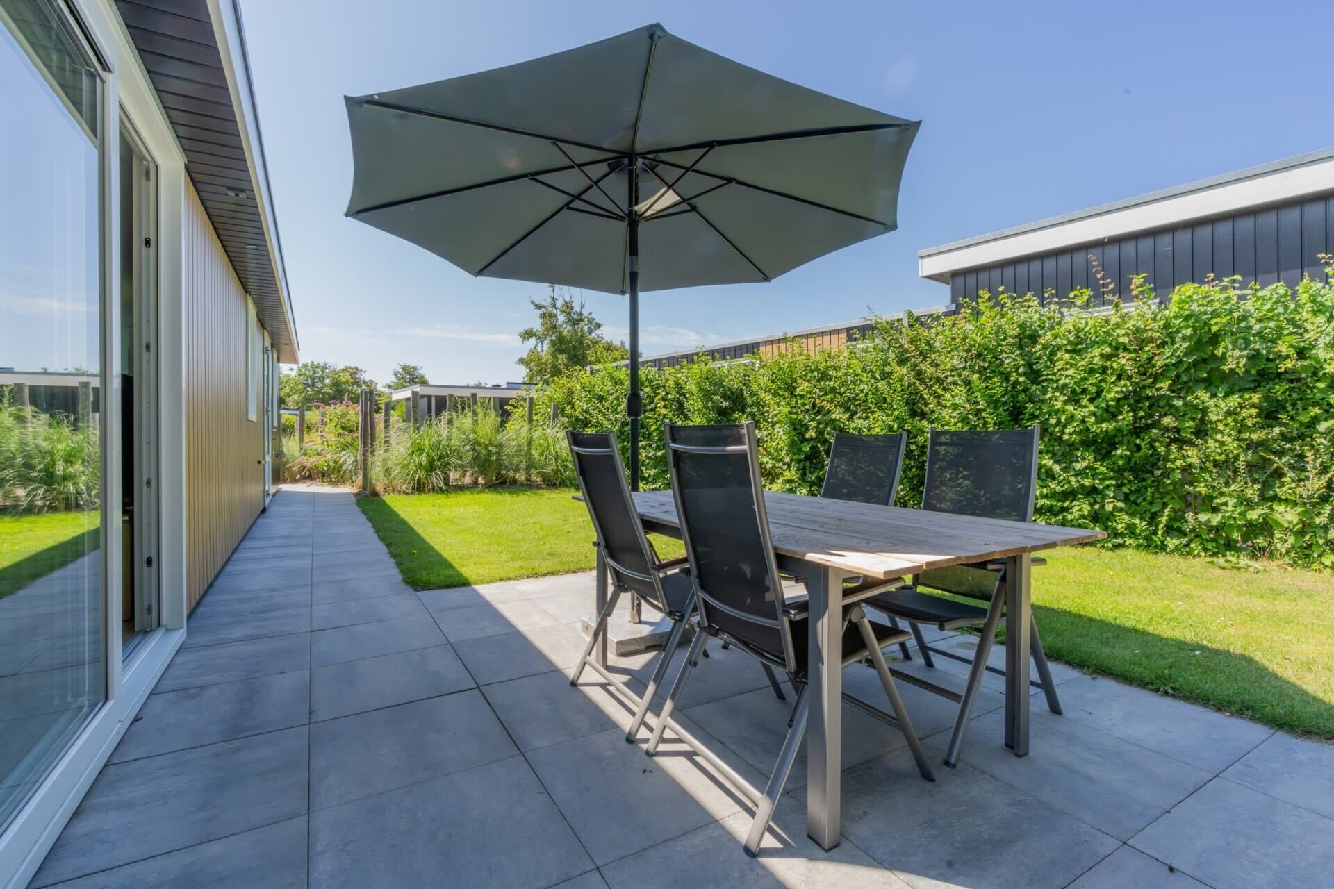 An outdoor patio with a table and chairs, a large umbrella, and a view of a modern building and greenery.