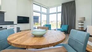 A modern living room with a round wooden table, blue sofas, a large window with curtains, and a flat-screen TV. - This modern chalet with 'Beach style' finish is located on a spacious plot and suitable for 4 people (Burgh-Haamstede)