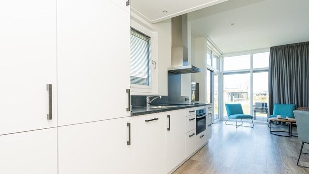 A modern kitchen with white cabinets, a black countertop, and a window with blinds. There is a dining area with a blue chair and a table. - This modern chalet with 'Beach style' finish is located on a spacious plot and suitable for 4 people (Burgh-Haamstede)