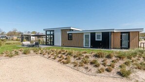 A modern two-story house with a flat roof, large windows, and a covered outdoor seating area. - This modern chalet with 'Beach style' finish is located on a spacious plot and suitable for 4 people (Burgh-Haamstede)