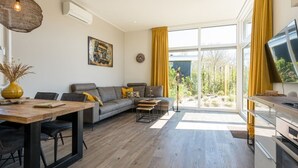 Ein modernes Wohnzimmer mit Sofa, Couchtisch und Esstisch. Ein großes Fenster mit Vorhängen, ein Gemälde an der Wand und ein Fernseher auf der rechten Seite.