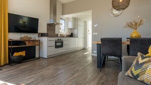 Eine moderne Küche mit weißen Schränken, ein Essbereich mit Tisch und Stühlen sowie ein Wohnzimmer mit Sofa und Fernseher.