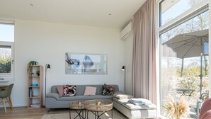 A modern living room with a grey sofa, a coffee table, and a large window with curtains. - This attractive chalet is located on a spacious plot and is suitable for 4 people. The garden, laid (Burgh-Haamstede)