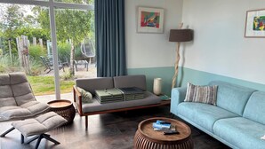 Un salon moderne avec un canapé, un fauteuil et une table basse, une grande fenêtre avec des rideaux et deux œuvres d’art encadrées accrochées au mur.