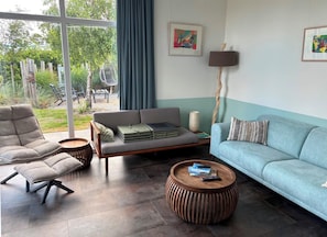 Een moderne woonkamer met een bank, fauteuil en salontafel, een groot raam met gordijnen, en twee ingelijste kunstwerken aan de muur.