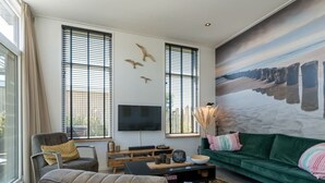 Una sala moderna con un sofá verde, un sillón marrón y una mesa de centro negra. En la pared, un cuadro grande representa una escena de playa.