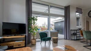A modern living room with a flat-screen TV, comfortable seating, a large sliding glass door leading to an outdoor area, and a dining area with a table and chairs.