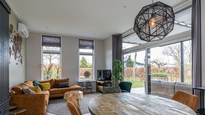 A modern living room with a round wooden dining table, leather sofas, and a large window with a view of a garden.