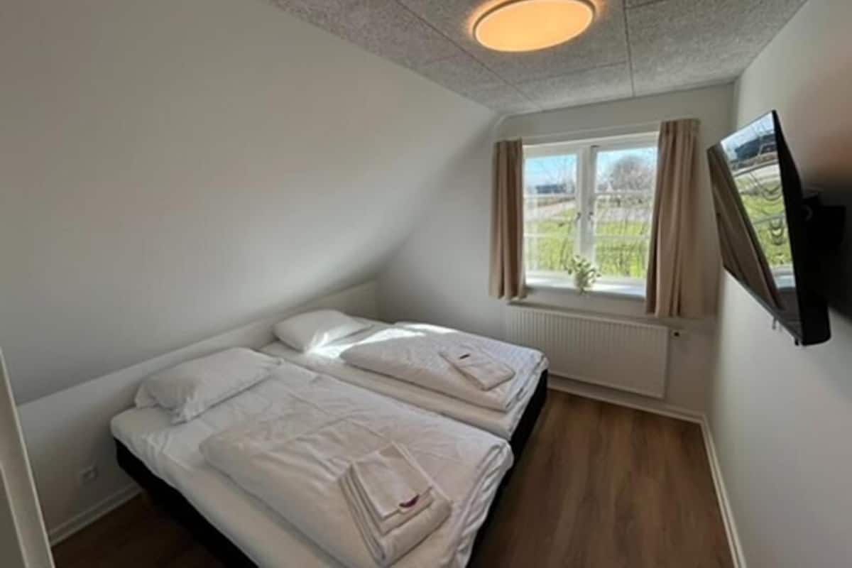 A small, clean room with two beds, a window with curtains, a mounted TV, and a ceiling light.