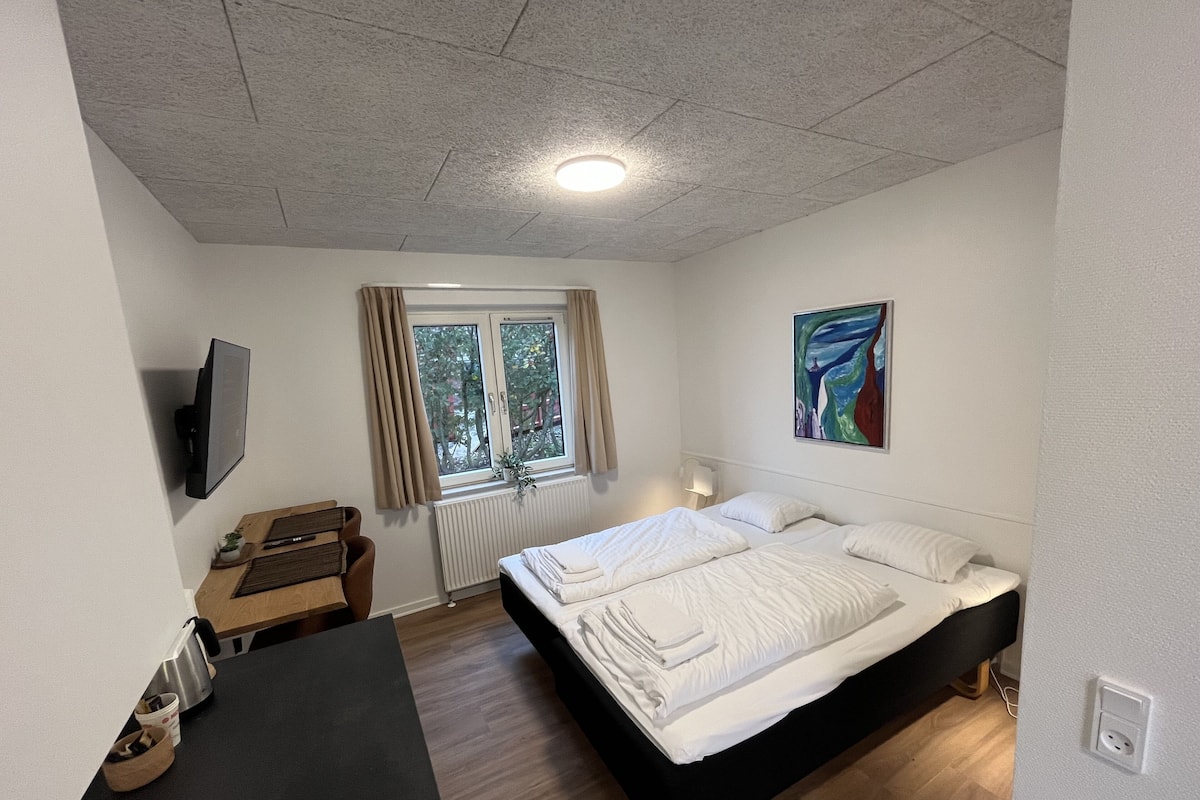A bedroom with a bed, a desk, a chair, a TV, a window, and a painting.