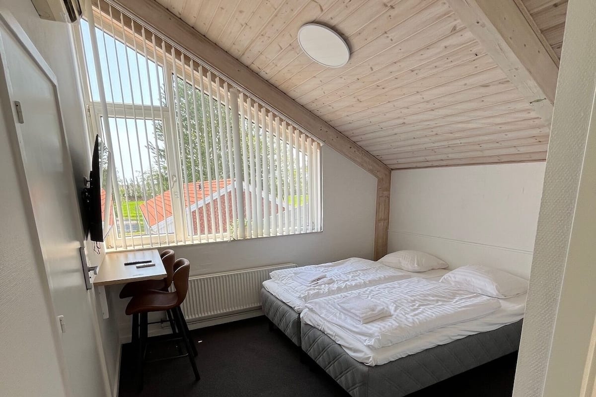A small, single-room accommodation with a sloped ceiling, a bed, a desk with a chair, and a window with blinds.