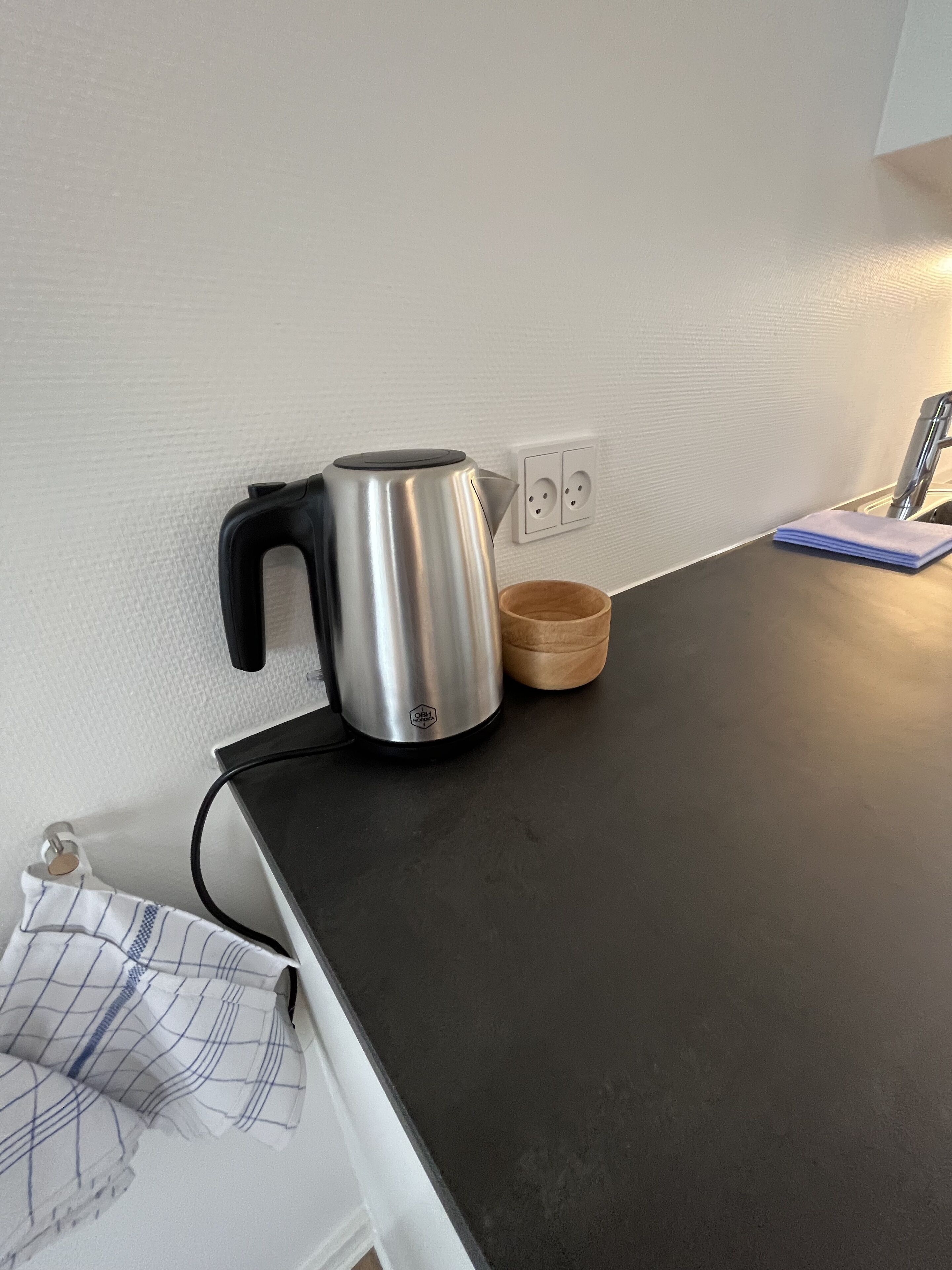 Une bouilloire en acier inoxydable est posée sur un plan de travail foncé, à côté d’un bol en bois. Une prise de courant et un linge plié se trouvent à proximité.