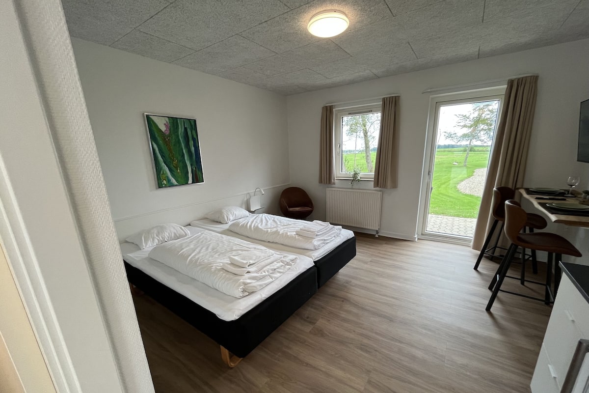 A bedroom with a bed, a desk, and a chair. There is a window with curtains and a view of the outdoors.
