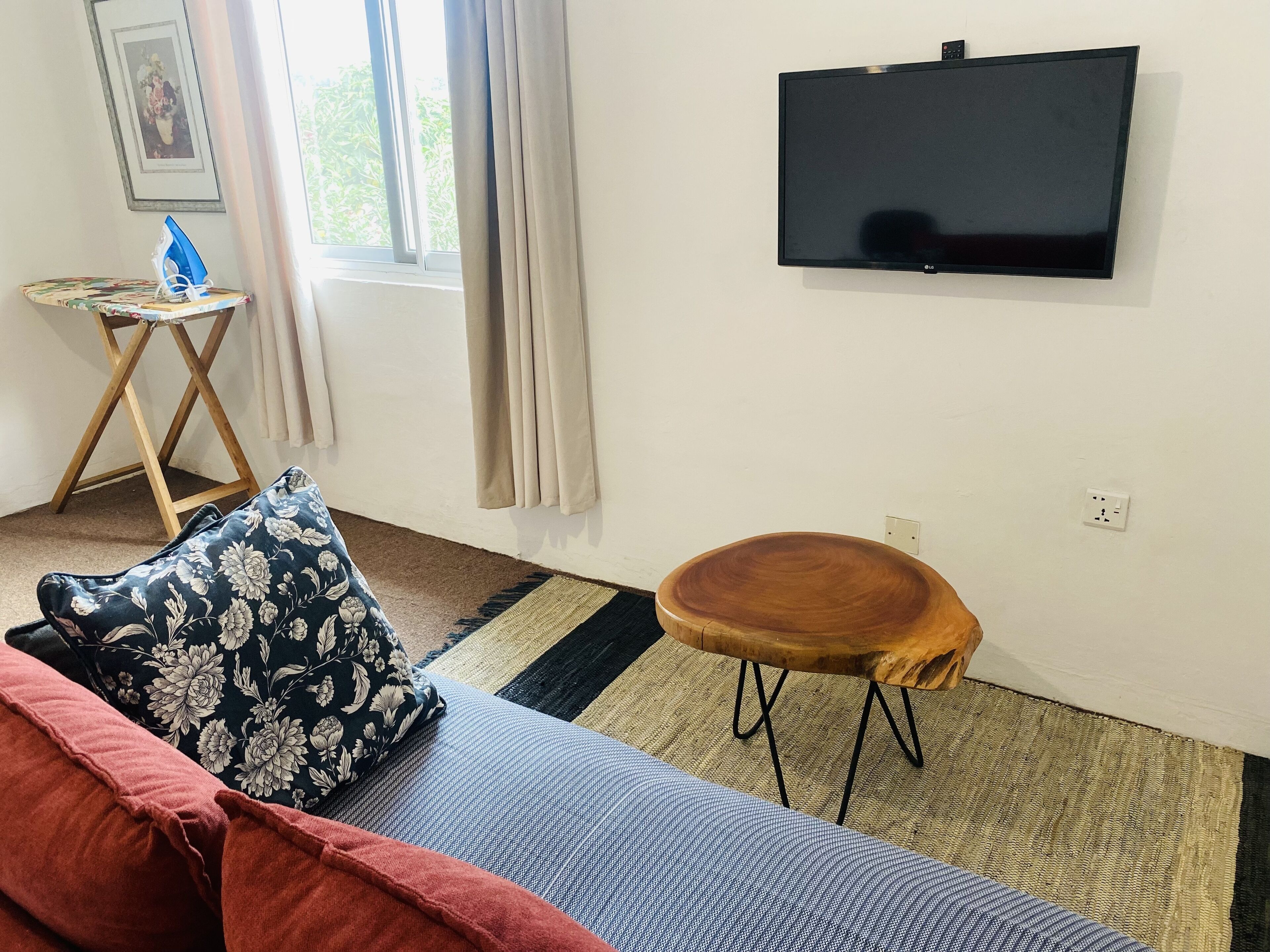 Ein Zimmer mit einer Couch, einem kleinen Holzschemel, einem an der Wand befestigten Fernseher und einem Fenster mit Vorhängen.