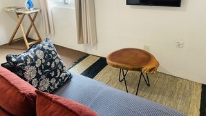 Ein Zimmer mit einer Couch, einem kleinen Holzschemel, einem an der Wand befestigten Fernseher und einem Fenster mit Vorhängen.