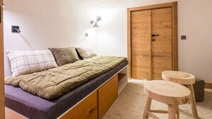 Quarto com uma cama, guarda-roupa de madeira, um banquinho pequeno e uma luminária fixada na parede.