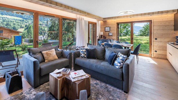 Una sala moderna con un sofá grande, una mesita de centro y un comedor con mesa y sillas. Amplios ventanales ofrecen vistas al exterior.