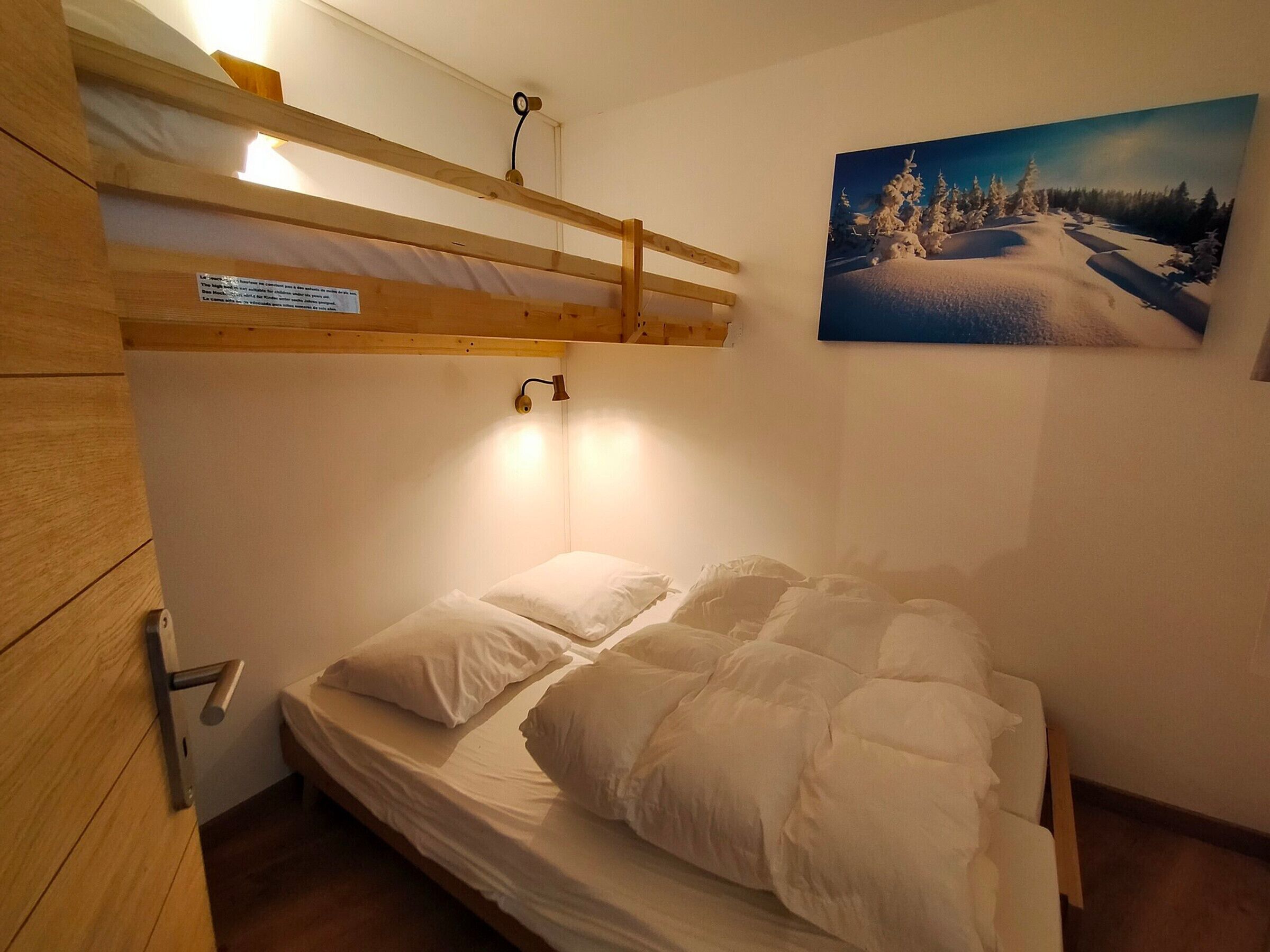 Un lit superposé avec une tête de lit en bois, un lit simple en dessous, une lampe fixée au mur et un tableau représentant un paysage enneigé.