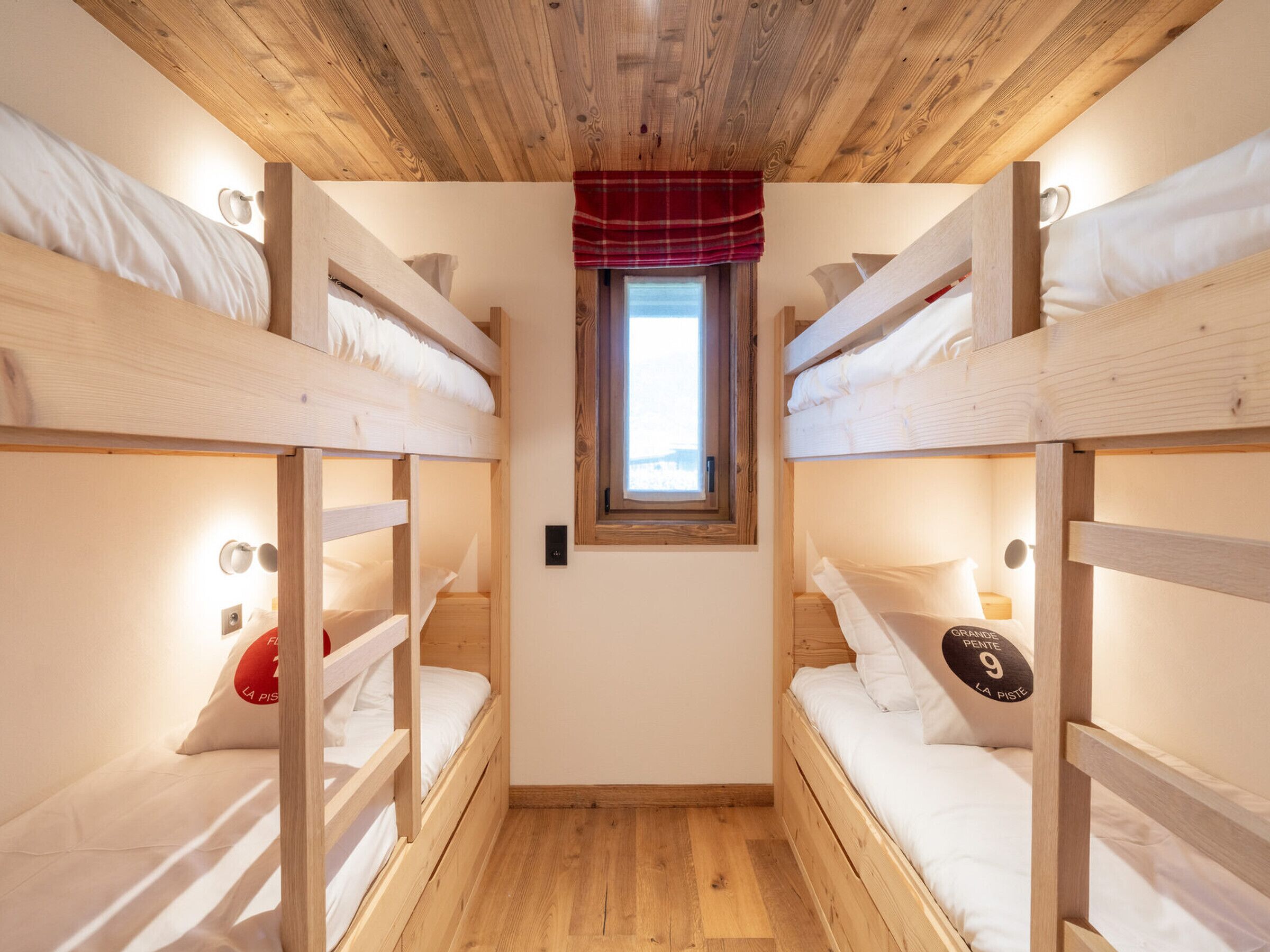 A room with bunk beds, a small window with a red valance, and a wooden ceiling.