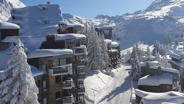 A snow-covered mountain village with multi-story buildings, a ski slope, and snow-covered trees.