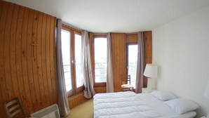A room with a large bed, a chair, a small table, and a window with curtains.