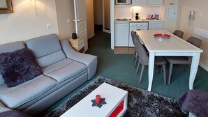En kompakt opholdsstue med sofa, spisebord, et køkken med hårde hvidevarer, og en gang der fører til et andet rum.