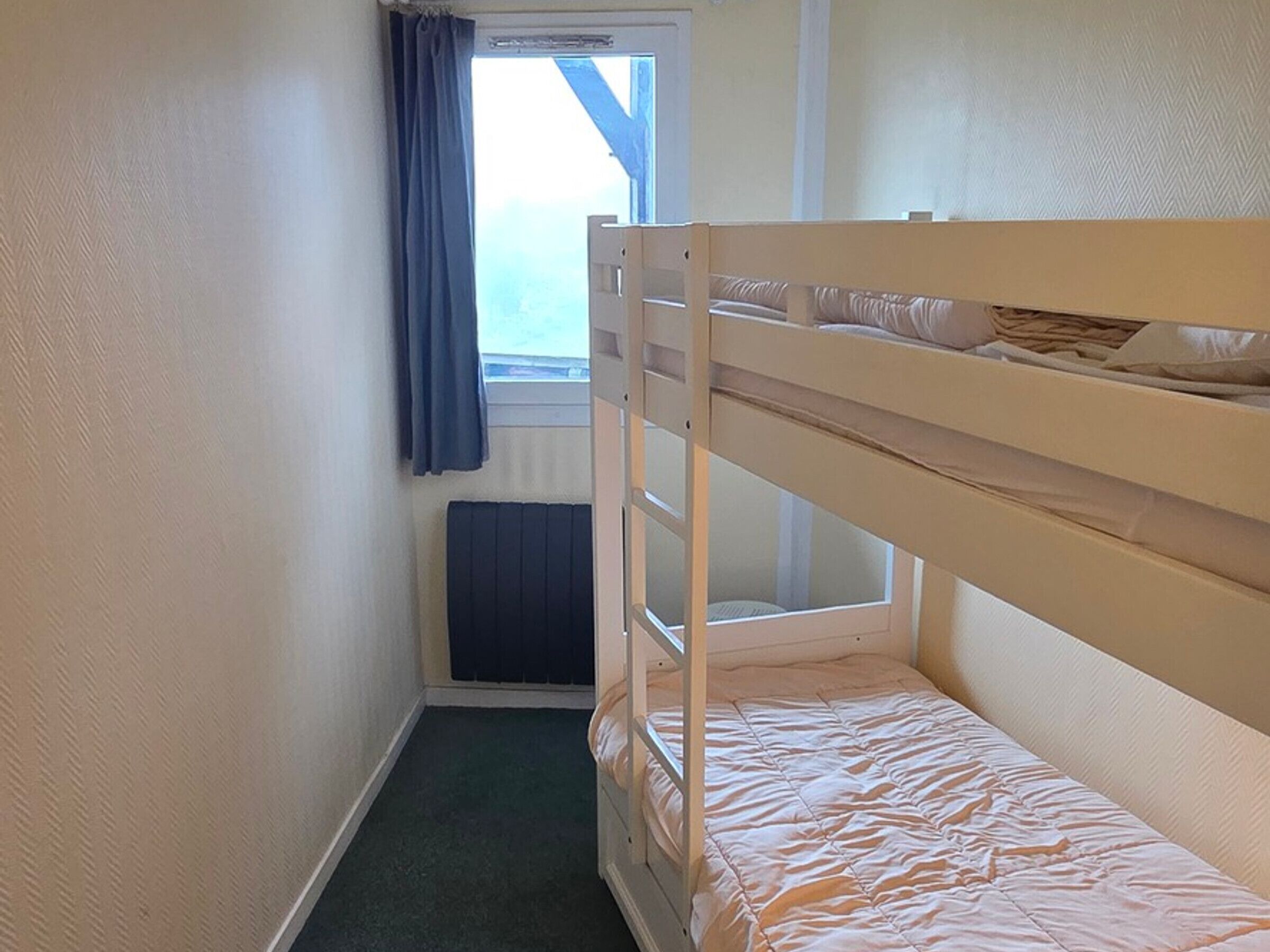A small, single-bunk room with a window, a radiator, and a door.