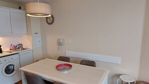 Et lille køkken med vaskemaskine, et bord med stole, et ur og en telefon på væggen.