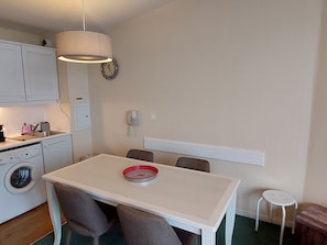 A compact kitchen with a washing machine, a small table with chairs, a clock, and a phone on the wall.