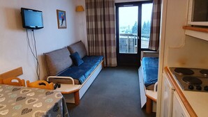 A compact hotel room with a sofa bed, a small kitchenette, a television, and a window with curtains.