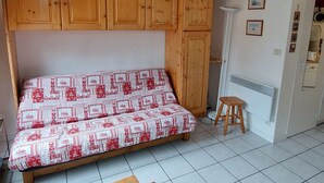 A small, cozy room with a wooden cabinet above a patterned sofa, a stool, and a radiator.