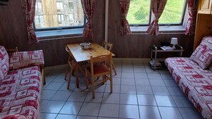 A compact living space with a dining table, two sofas, and two windows with curtains.