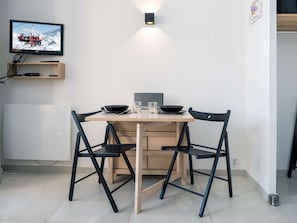 Quarto pequeno e moderno com uma mesa de madeira e duas cadeiras pretas, uma TV de tela plana na parede e uma pequena prateleira abaixo.