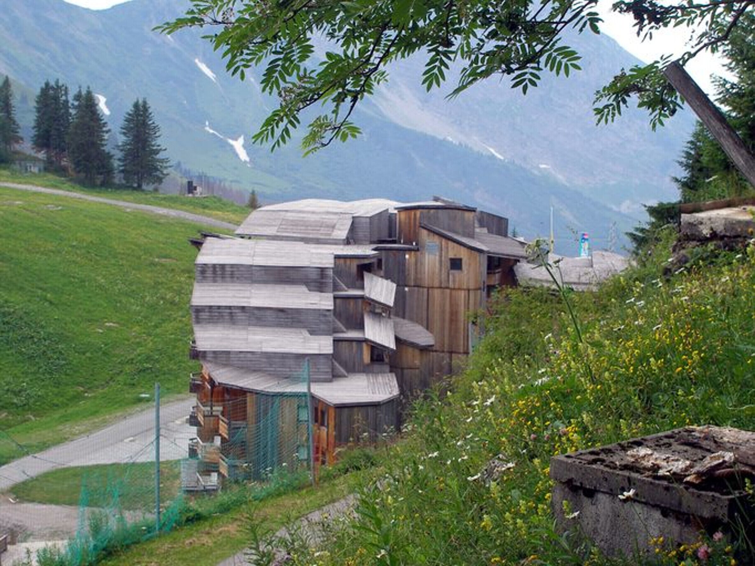 Un edificio de madera rústico con varios balcones, rodeado de un paisaje verde exuberante y montañas al fondo.