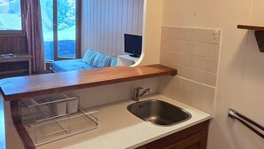 A compact kitchen with a sink, a drying rack, and a window overlooking a balcony with a bed and a TV.