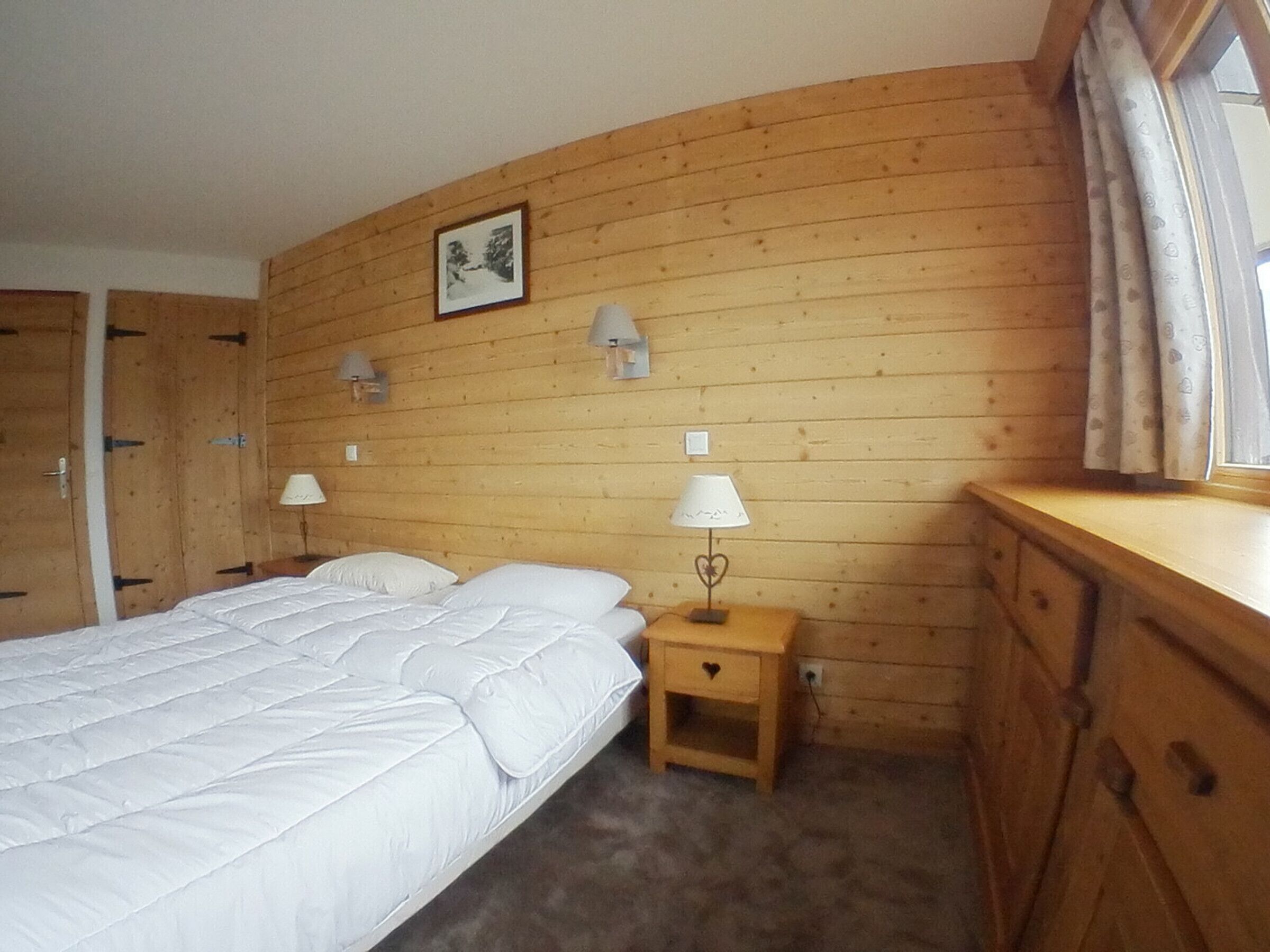A bedroom with a wooden headboard, a bed with white bedding, two bedside lamps, a small bedside table with a lamp, a picture on the wall, and a window with curtains.