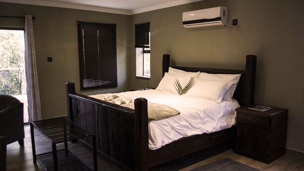 Una cama bien hecha con estructura de madera y ropa blanca, una mesita de noche pequeña, una mesita de noche oscura, una ventana con persianas y un equipo de aire acondicionado en la pared.