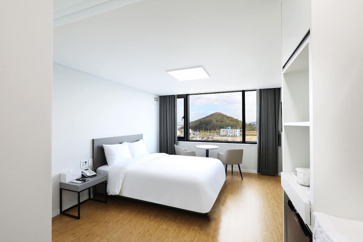 Una habitación de hotel con cama, mesita de noche, una pequeña mesa con sillas, una ventana con vistas a una colina y edificios, y un minibar.
