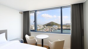 Habitación de hotel con cama, dos sillas, una mesita y un ventanal grande con vistas al puerto y a una colina.