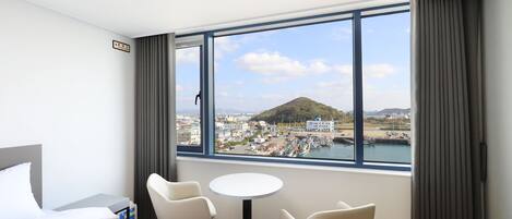 Habitación de hotel con cama, dos sillas, una mesita y un ventanal grande con vistas al puerto y a una colina.