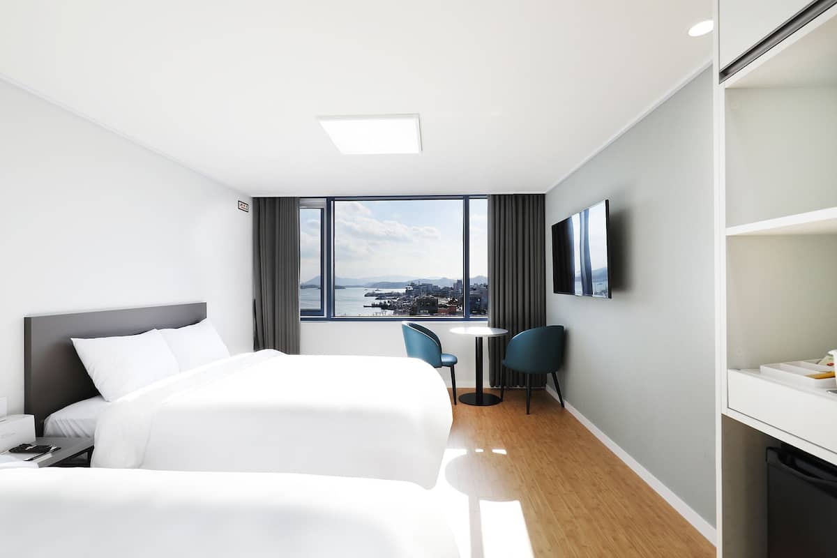 Habitación de hotel con dos camas, un escritorio, una silla, un televisor y un ventanal con vistas a la ciudad.